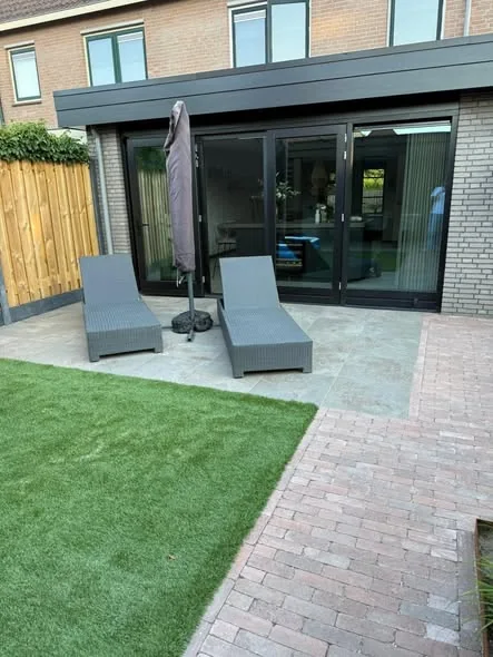 Terras van grote tegels bij de achtergevel, met een pad van gebakken klinkers en kunstgras