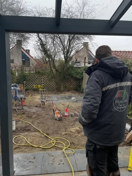 Mick Wiemerink op de rug gezien in een werkjas met het logo, kijkend naar een afgegraven tuin waar de eerste tegels liggen