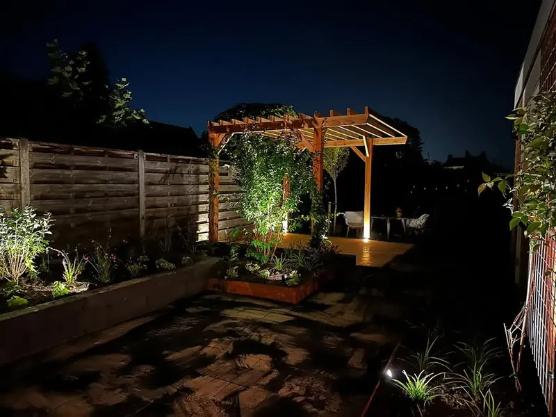 Tuin bij avond met verlichte borders, een verhoogde border en een houten pergola