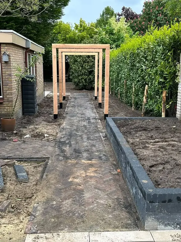 Pad in keperverband onder een rij houten pergolabogen, naast een verhoogde border van donkere stenen