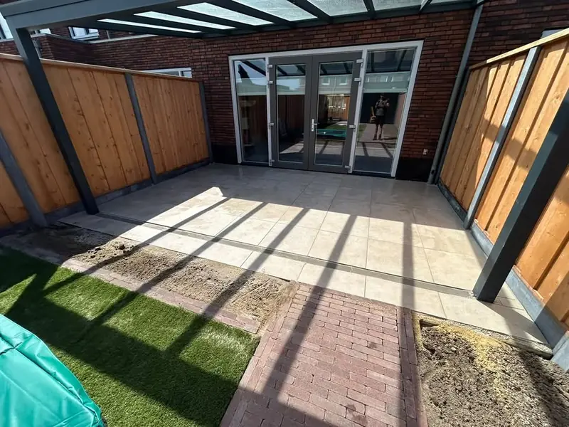 Terras van lichte tegels onder een glazen overkapping, met een lijngoot, een pad van klinkers en kunstgras