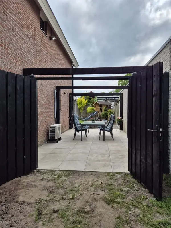 Zwarte houten poort met balken erboven, met daarachter een terras van lichte tegels en een tuintafel